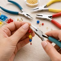 Close-up of hands using rubber-tipped flat nose jewelry pliers to assemble a delicate beaded pendant, with assorted jewelry pliers, wire, and findings in the background.