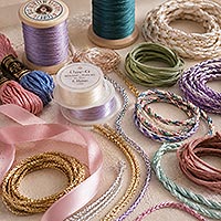 Assorted jewelry and craft cords including satin ribbon, braided cords, metallic cords, suede lace, embroidery floss, beading thread, and decorative string arranged in soft pastel colors.