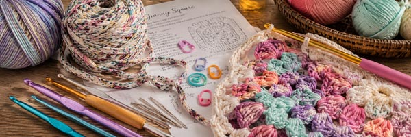 Bright crochet crafting scene with colorful yarn, fabric strip yarn, crochet hooks, stitch markers, wide-eye tapestry needles, a crochet pattern, and a partially completed crochet project displayed on a wooden tabletop.